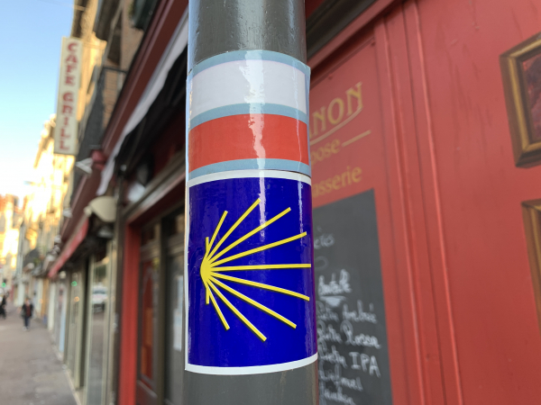 Rue Saint-Jacques, en face de l'église, nous sommes sur la bonne route ! La fameuse coquille jaune stylisée sur fond bleu nous rappellera que nous sommes sur un chemin un peu spécial. Mais cette balise est peu fréquente, c'est surtout le balisage rouge et blanc du GR210 qui nous guidera.
